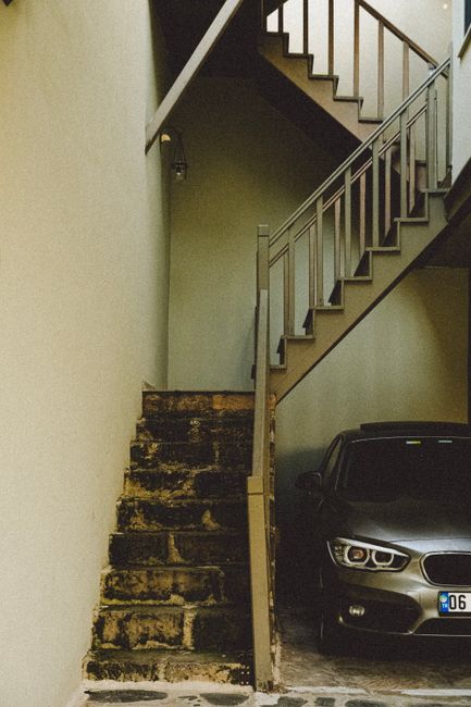 A narrow indoor stairwell with a set of old, worn concrete stairs on the left, showing signs of dirt and damage, leading up to a higher floor. To the right of the stairs, there is a modern metal staircase with wooden handrails, ascending in the opposite direction. Below the staircase, a black vehicle is parked inside a parking area adjacent to the stairwell, partially visible with a contemporary design. The surrounding walls are painted in neutral tones, with a small wall-mounted light fixture visible near the top left corner. The lighting is soft, primarily coming from above, illuminating the entryway during a home relocation process. The scene reflects the typical environment encountered during house removals where space constraints and structural elements may present access challenges that Man with Van Kingston addresses in their services for flat removals, particularly in locations such as Kingston with stair and parking issues.