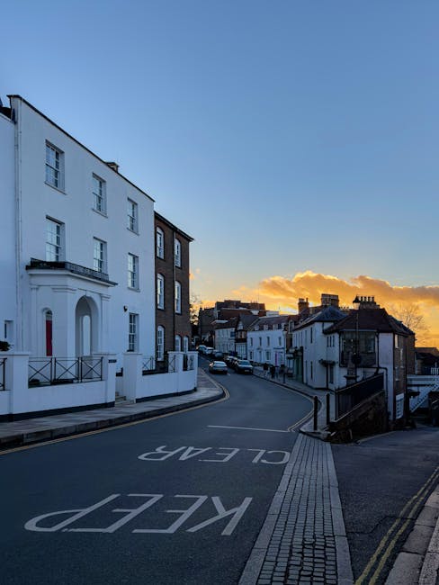 A narrow residential street in Kingston during sunset, featuring a mix of white and dark brick terraced houses and apartment buildings on either side. The street has a gentle upward slope with a clearly marked 'KEEP CLEAR' road sign painted on the asphalt. On the left, there is a large white building with multiple windows, decorative balconies, and a small front garden enclosed by a black metal fence. Adjacent to it, a dark brick building with large windows can be seen. The pavement on both sides includes tactile paving for pedestrians. The sky displays warm orange and yellow hues with some clouds near the horizon, creating a peaceful evening atmosphere. This scene illustrates typical urban residential surroundings in Kingston, suitable for a discussion on house removals or relocation planning, especially considering access challenges on steep or narrow streets as handled by companies like Man with Van Kingston.