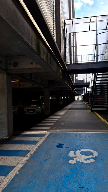 A narrow indoor stairwell with a set of old, worn concrete stairs on the left, showing signs of dirt and damage, leading up to a higher floor. To the right of the stairs, there is a modern metal staircase with wooden handrails, ascending in the opposite direction. Below the staircase, a black vehicle is parked inside a parking area adjacent to the stairwell, partially visible with a contemporary design. The surrounding walls are painted in neutral tones, with a small wall-mounted light fixture visible near the top left corner. The lighting is soft, primarily coming from above, illuminating the entryway during a home relocation process. The scene reflects the typical environment encountered during house removals where space constraints and structural elements may present access challenges that Man with Van Kingston addresses in their services for flat removals, particularly in locations such as Kingston with stair and parking issues.
