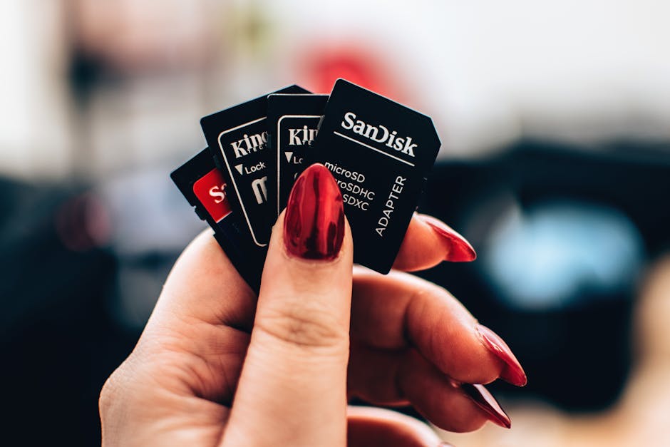 Close-up of a person's hand with long, polished red nails holding four SD memory cards, including brands like Kingston and SanDisk, with labels indicating microSDHC and SDXC formats. The background is blurred, suggesting an indoor setting, possibly related to packing or organizing electronic equipment during a house move. The memory cards are held between the thumb and index finger, with the other fingers slightly curved. This image illustrates aspects of packing, data transfer, or planning involved in home relocation or furniture transport, aligning with professional removals services such as those provided by Man with Van Kingston for efficient moving and packing processes.