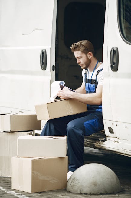 A man wearing a striped shirt and blue work overalls is sitting on the edge of an open white delivery van, holding a clipboard and writing notes. Around him are several cardboard boxes in various sizes, some stacked on the pavement and others nearby, ready for transport. The open van door reveals more packing materials inside, with a focus on the loading process during a home relocation. The scene is set outdoors, with concrete pavement and a concrete bollard visible in the foreground, emphasizing a professional furniture transport and packing and moving activity. Man with Van Kingston is providing a professional removals service, efficiently managing the logistics of house removals and furniture transport.