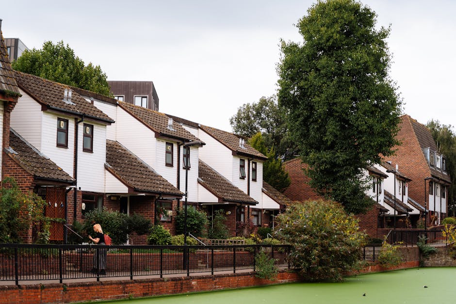 A row of modern multi-story houses with brick facades and red-tiled roofs situated along a canal, reflecting on the calm water surface below. Each property features small balconies with black metal railings, white-framed windows, and well-maintained landscaped gardens with bushes and purple flowering plants. A white motorboat is docked at a wooden pier adjacent to one of the houses. The scene is captured during sunset, with a colorful sky overhead. In the foreground, the canal’s water provides a clear mirror image of the houses, boat, and surrounding greenery. This setting illustrates a peaceful riverside residential area that could benefit from home relocation and furniture transport services, as provided by Man with Van Kingston for house removals and moving logistics.