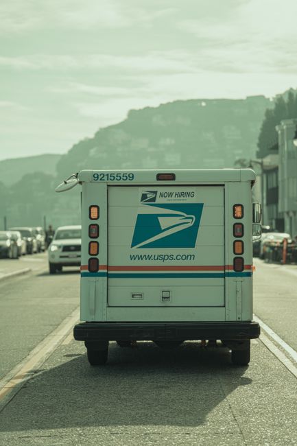 A white USPS delivery van with the USPS logo and the text 'NOW HIRING' on its rear doors, parked on a street with several vehicles visible on either side. The van is positioned close to the curb, and