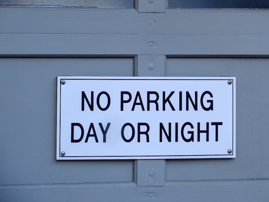 Close-up view of a rectangular white metal sign mounted on a light grey wooden surface, featuring bold black text that reads 'NO PARKING DAY OR NIGHT'. The sign is secured with four screws, one in each corner. The background shows the textured wooden paneling with visible horizontal and vertical lines, likely part of a garage or warehouse exterior. This image relates to Kingston Council permit rules for furniture removals and parking, illustrating restrictions that may impact home relocation or furniture transport processes, as managed by Man with Van Kingston. The lighting is neutral, highlighting the sign's clarity and the surface details around it.