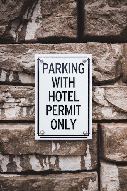 A rectangular metal sign attached to a rough stone wall, displaying the message 'PARKING WITH HOTEL PERMIT ONLY' in bold black letters on a white background. The sign is fixed with four screws, one in each corner. In the context of house removals or furniture transport, this sign indicates restricted parking requirements for vehicles involved in a property relocation. The wall behind the sign consists of large, irregularly shaped stones with visible mortar, and the lighting is natural, highlighting the texture of the stone surface. The image is relevant to Kingston Council permit rules for furniture removals and parking, as managed by Man with Van Kingston, emphasizing the importance of adhering to parking regulations during home relocation and moving processes.