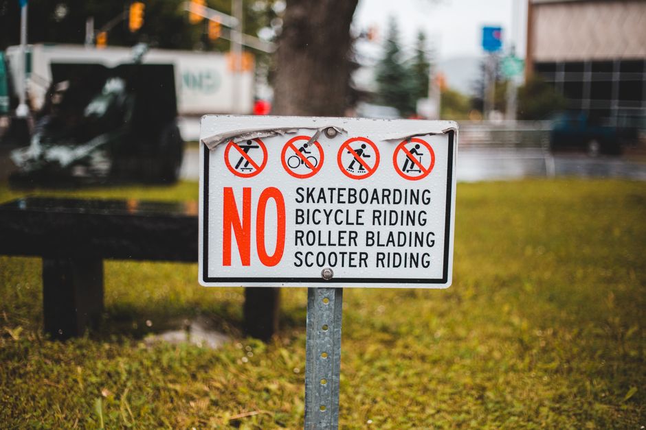 Close-up view of a rectangular white metal sign mounted on a light grey wooden surface, featuring bold black text that reads 'NO PARKING DAY OR NIGHT'. The sign is secured with four screws, one in each corner. The background shows the textured wooden paneling with visible horizontal and vertical lines, likely part of a garage or warehouse exterior. This image relates to Kingston Council permit rules for furniture removals and parking, illustrating restrictions that may impact home relocation or furniture transport processes, as managed by Man with Van Kingston. The lighting is neutral, highlighting the sign's clarity and the surface details around it.