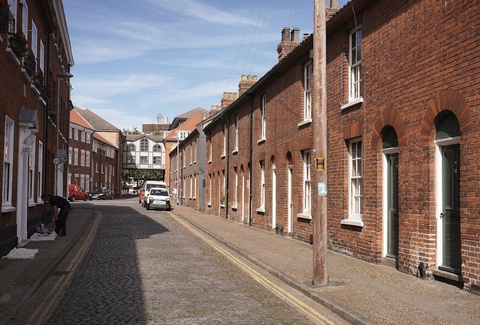 A narrow cobblestone street in Kingston lined with a series of red-brick terraced houses, each featuring white-framed sash windows and white doors, with some doors open during daylight hours. On the left side of the street, a person appears to be wrapping or packing items on the pavement, possibly preparing for a home relocation. A black trolley is visible nearby, along with cardboard boxes and packing materials spread on the sidewalk. Several vehicles, including a white van and a red car, are parked along the curb, with the van positioned close to the entrance of one of the terraced houses where loading or unloading may be taking place. A wooden utility pole is situated near the curb, with overhead cables extending across the street. In the background, a modern, multi-storey building with large windows and a bridge-like structure connects two sections of the development, under a clear blue sky with some wispy clouds, highlighting a typical urban scene involving furniture transport and packing during a house removals process in Kingston, facilitated by Man with Van Kingston.