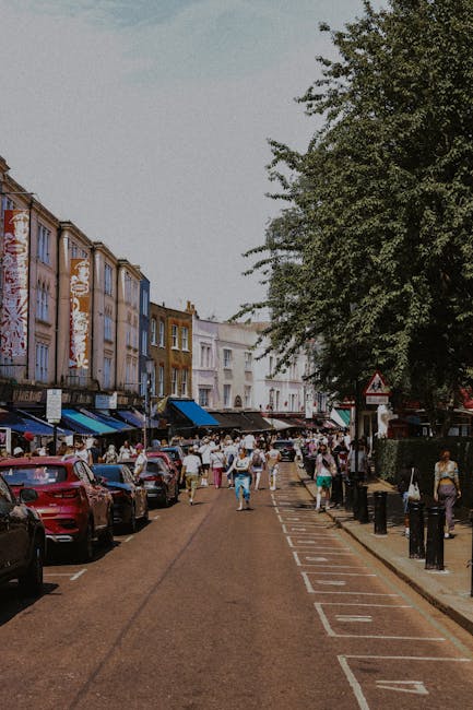 A busy street scene in Kingston with a row of multistory buildings on the left side, featuring shops and cafes with awnings and signage. Parked cars line the curb along the street, with several pedestrians walking on the pavement, some carrying boxes or bags. On the right side, there is a designated parking area marked with white lines and black bollards, adjacent to a large, leafy tree providing shade. In the background, more buildings and trees are visible under a partly cloudy sky, indicating daytime. The scene captures elements typical of urban house removals and furniture transport, including the presence of a moving van or vehicle possibly used by Man with Van Kingston for home relocation services, with objects like cardboard boxes and wrapped furniture nearby. Overall, the image conveys the active logistics associated with packing, loading, and moving process in a residential and commercial setting.