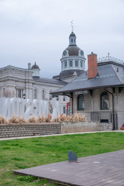 The image shows an exterior view of a historic building with a prominent domed clock tower featuring a weather vane on top, situated in Kingston. In the foreground, there is a well-maintained green la