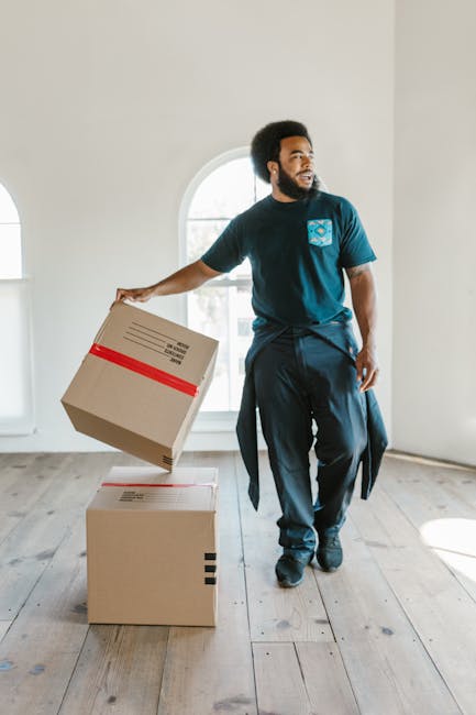 A man with dark curly hair and a beard, wearing a blue T-shirt with a logo on the chest and dark trousers, is inside a bright, spacious room with white walls and wooden flooring. He is lifting a cardboard moving box with red tape around it, preparing to load it onto a lower box that is already placed on the floor. The room features a large, arched window letting in natural light, indicating daytime. The scene depicts the process of home relocation, with the individual handling packing and furniture transport tasks as part of a professional removals service, such as those provided by Man with Van Kingston, emphasizing safe packing and organized loading within a residential setting.