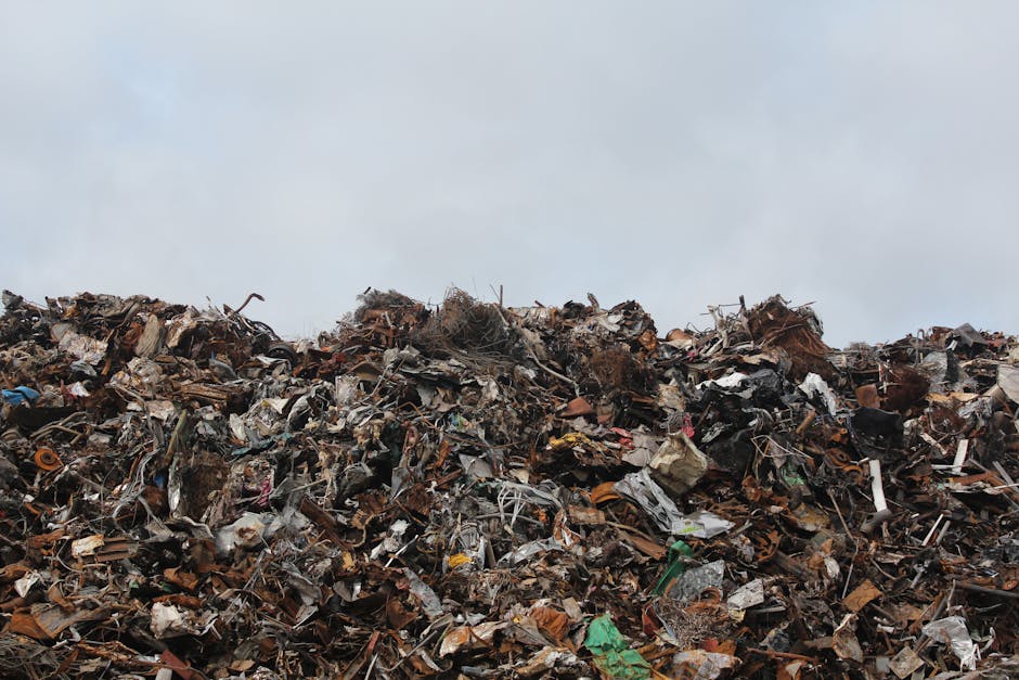 A red metal communal waste skip with visible rust and scratches is positioned against the corner of a building exterior, on a paved sidewalk area. Next to the skip, several tied plastic garbage bags f